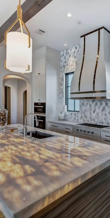 Backlit onyx island countertop beside a white and gold range hood and stainless wall ovens