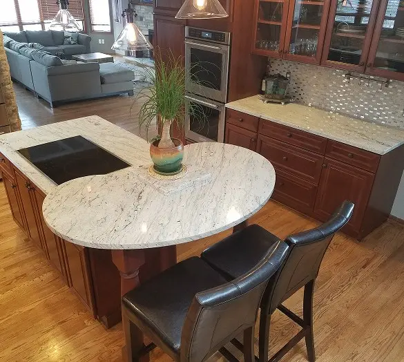 Curved granite kitchen island with a cooktop and bar seating, surrounded by cherry cabinetry, wall ovens, and a mosaic tile backsplash