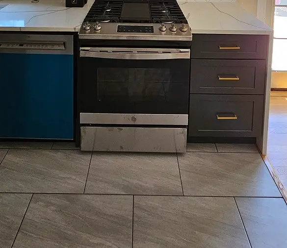 Gray porcelain tile kitchen flooring beside a stainless steel slide-in range, dark cabinets with brass pulls, and white quartz countertops
