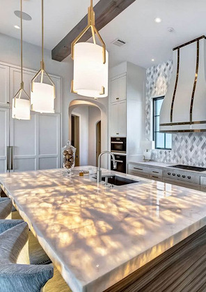 Large kitchen island with a softly veined white stone top under gold-framed pendant lights, with a brass-strapped range hood