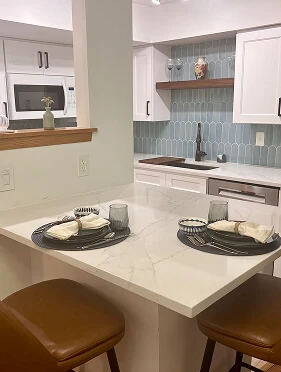 White kitchen peninsula with quartz countertop, blue picket tile backsplash, floating wood shelf, and leather barstools