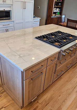 White kitchen island with a light veined stone countertop and a black gas cooktop, beside a wood display shelf