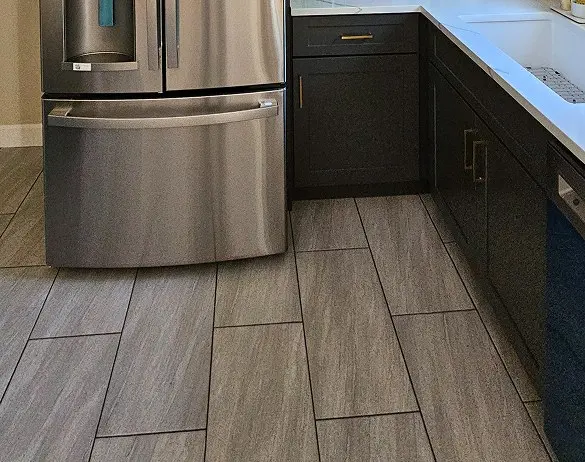 Gray wood-look tile flooring in a kitchen with dark espresso cabinets, brass hardware, white countertops, and a stainless steel refrigerator